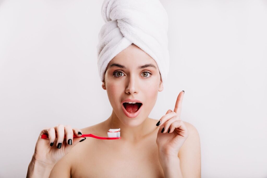 Млада жена с хавлиена кърпа на главата е вдигнала предуредително пръст и се готви да измие зъбите си с четка и паста за зъби.